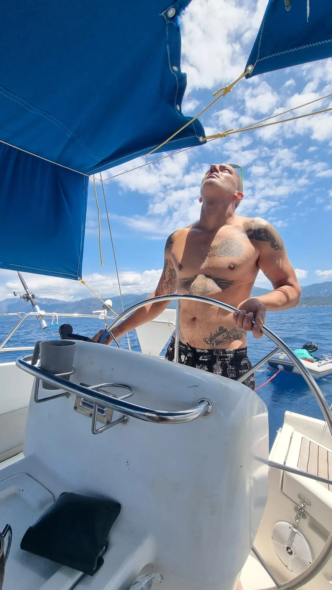 Stan, skipper, à la barre du voilier Eridu sous le taud bleu, montagnes corses en arrière-plan